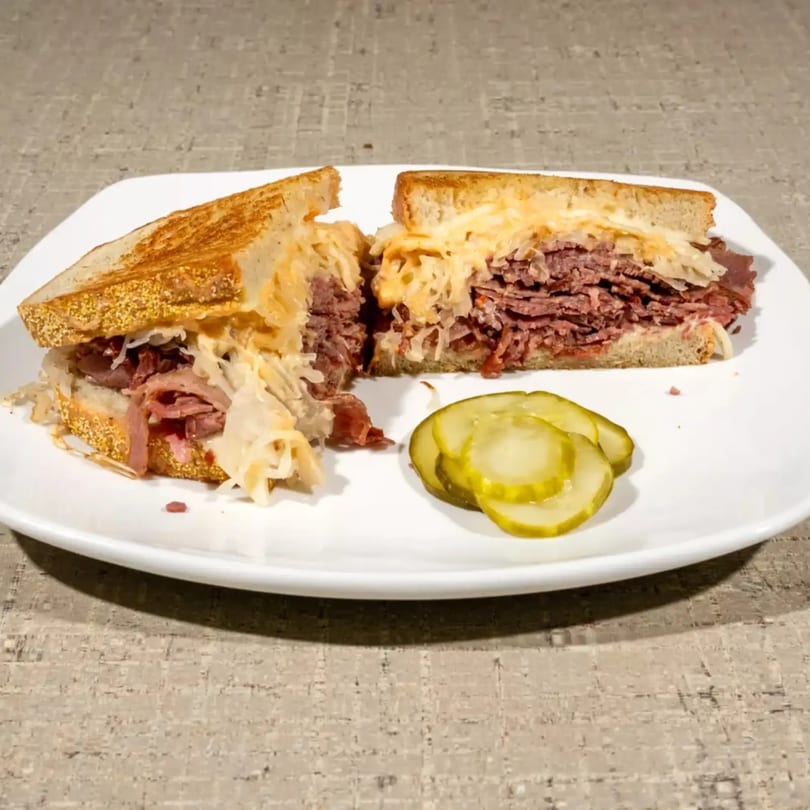 A grilled Reuben sandwich, cut in half, with pastrami, sauerkraut, and cheese, served with pickles.
