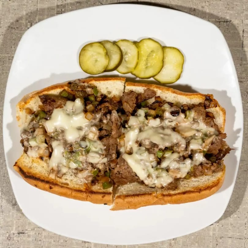 Cheesesteak sandwich with melted cheese, peppers, and onions, served with pickle slices on a white plate.