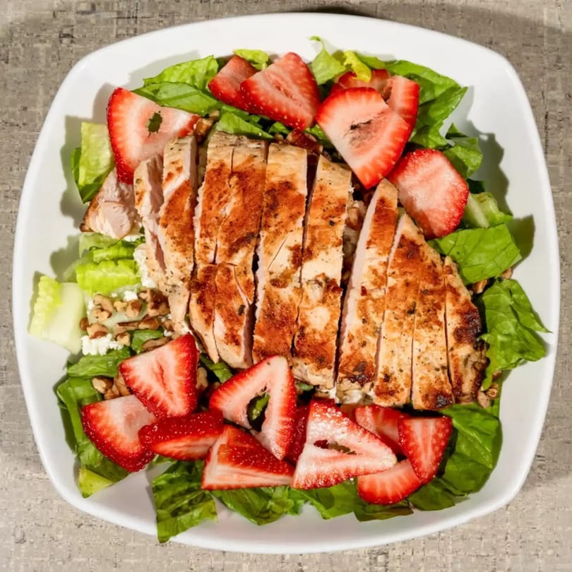 Overhead view of a salad with sliced grilled chicken, strawberries, lettuce, feta, and nuts.