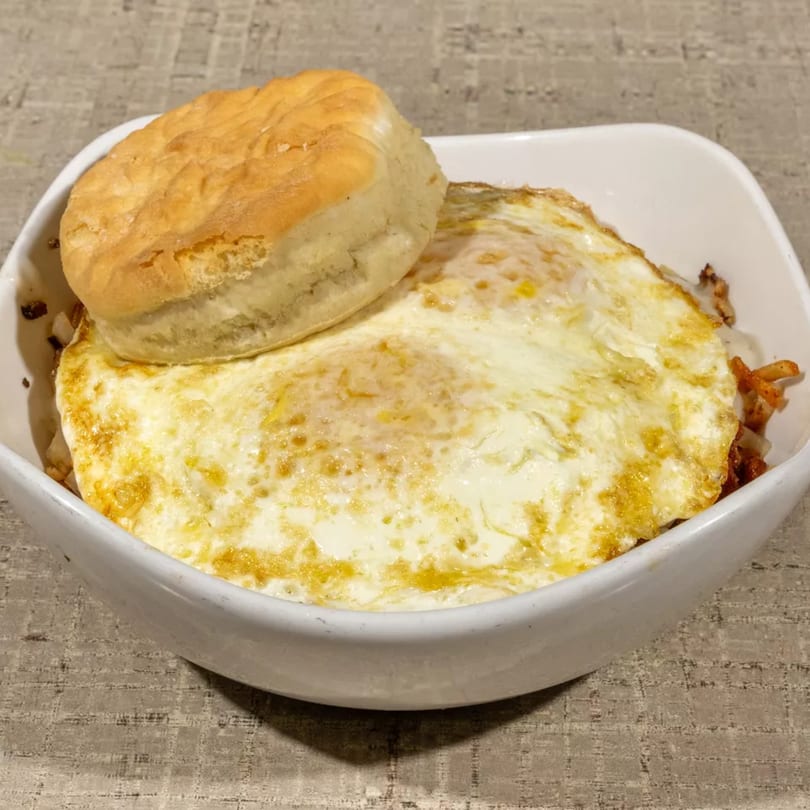 A white bowl filled with fried eggs and hash browns, topped with a golden biscuit.